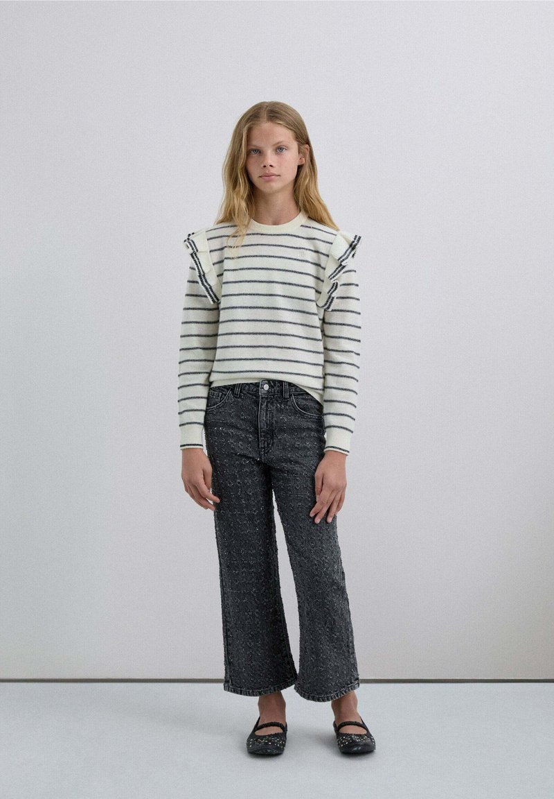 Randig tröja med volangdetaljer på axlarna, kombinerad med strukturerade svarta vida jeans och platta skor med utsmyckningar.