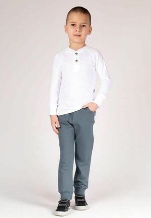 Biała koszula z długim rękawem z plisą na trzy guziki, zestawiona z szarymi spodniami i czarnymi trampkami. Prosty fason, gładki materiał.