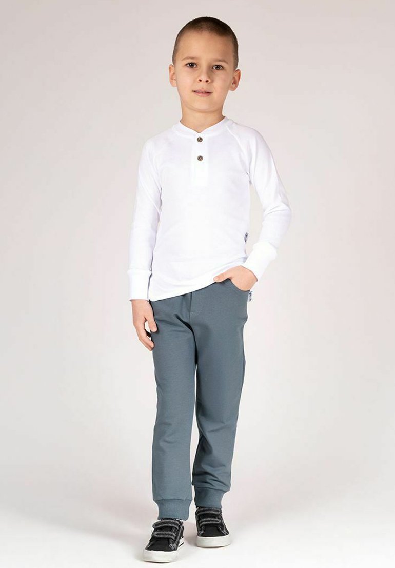 Biała koszula z długim rękawem z plisą na trzy guziki, zestawiona z szarymi spodniami i czarnymi trampkami. Prosty fason, gładki materiał.