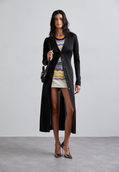 Larga chaqueta negra con botones, llevada sobre un vestido de estampado multicolor. La modelo sostiene un pequeño bolso negro y lleva zapatos de tacón brillantes.