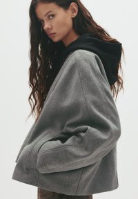 Jachetă oversized gri cu glugă, cu o textură moale și buzunare laterale. Designul este modern, cu o croială lejeră și cusături minime.