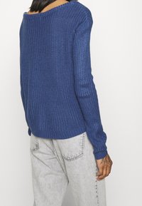 Pull bleu en tricot avec un large col, texture côtelée et manches longues, associé à un jean gris clair. Les caractéristiques notables incluent un motif en grosse maille.