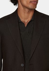 Blazer in lino marrone con rever a picco, chiusura a un bottone e una tasca piccola. Indossato sopra un polo lavorato a maglia scura con texture a rombi.