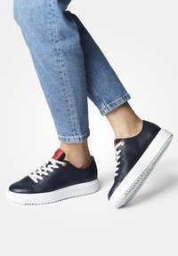 Baskets en cuir bleu marine avec semelle et lacets blancs, accents rouges au talon et finition texturée ; associées à un jean court bleu clair.