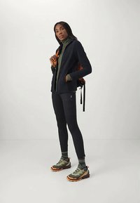 Tumesinine tõmblukuga jakk rohelise kõrge kaelusega, mustad legginsid, rohelised ja oranžid matkajalatsid ning oranžide aktsentidega seljakott.