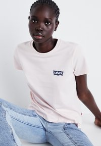 T-shirt rose clair en coton, arborant un logo Levi's bleu sur la poitrine. Porté avec un jean bleu clair, légèrement déchiré aux genoux.