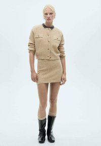 Beige stickad cardigan med framknappar och fickor, matchad med en minikjol. Avslutad med svarta knähöga stövlar och krämfärgade strumpor.