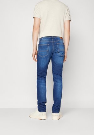 Personne portant un jean bleu, des baskets blanches et une chemise beige clair à manches courtes, debout de dos devant un fond uni.