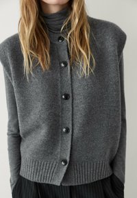 Harmaa neulecardigan, jossa on korkea kaulus, mustat napit ja ribbireunus. Materiaali näyttää pehmeältä ja siinä on teksturoitu pinta.