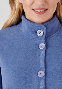 Chaqueta de forro polar en azul claro con un cuello alto, cierre de botones y un patrón de hojas bordado en la parte delantera. Presenta dos botones grandes.