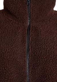 Braune Fleecejacke mit hohem Kragen und einem zentralen schwarzen Reißverschluss. Hat eine strukturierte Oberfläche mit einem weichen, flauschigen Aussehen.