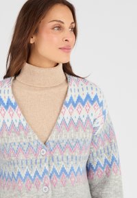 Cardigan grigio a fantasia con motivi a diamante rosa e blu, caratterizzato da un profondo scollo a V e due bottoni chiari. Indossato sopra un dolcevita beige.