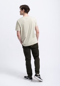 Lichtbeige katoenen t-shirt met korte mouwen en een subtiel logo op de achterkant, gecombineerd met donkergroene broek en zwarte sneakers.