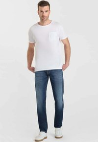 Camiseta blanca de manga corta con detalle de bolsillo texturizado, combinada con jeans de mezclilla oscura y zapatillas blancas con acentos en marrón claro.