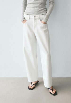 Personne portant un pantalon blanc à jambes larges, un haut gris à manches longues, des sandales noires type tongs, debout les mains dans les poches sur un sol gris.
