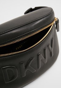 Sac bandoulière DKNY en cuir noir avec fermeture éclair dorée partiellement ouverte, logo en relief sur le devant et sangle ajustable visible.