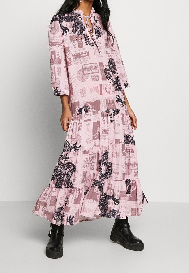 Růžové maxi šaty s dlouhým rukávem a stupňovitou spodní částí, s potiskem draka, textem a zavazováním na krku. Nosené s černými platformovými botami.