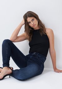 Top sin mangas negro acanalado combinado con jeans ajustados de cintura alta en azul oscuro. Modelo sentada, usando tacones negros con tiras, con un fondo neutro.