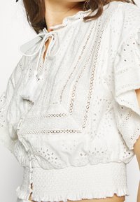 Weiße Baumwollbluse mit aufwendiger Stickerei, einem Rüschenausschnitt und gesmoktem Saum. Verstellbare Bänder und ausgestellte kurze Ärmel sorgen für visuelle Details.