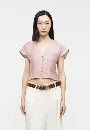 Jeune femme aux cheveux droits et foncés portant un crop top rose à manches courtes boutonné et un pantalon blanc taille haute avec une ceinture marron.