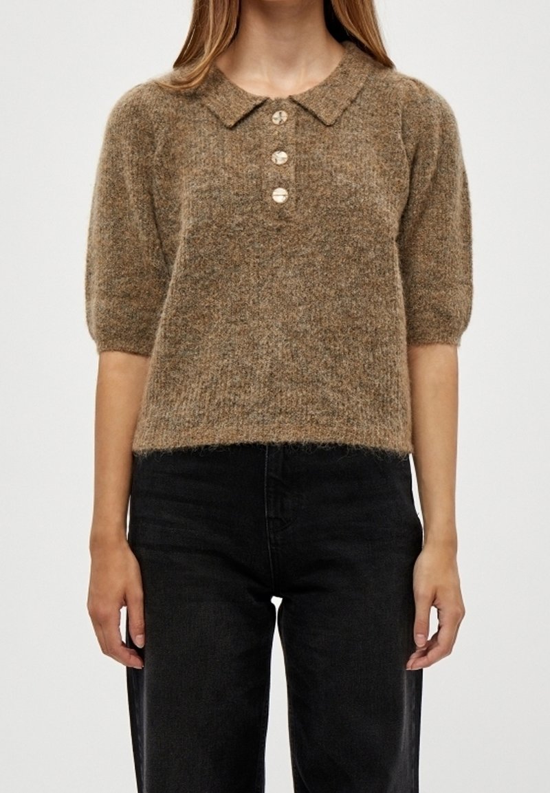 Femme portant un pull marron duveteux à manches courtes avec col et trois boutons, associé à un pantalon noir taille haute sur un fond uni.