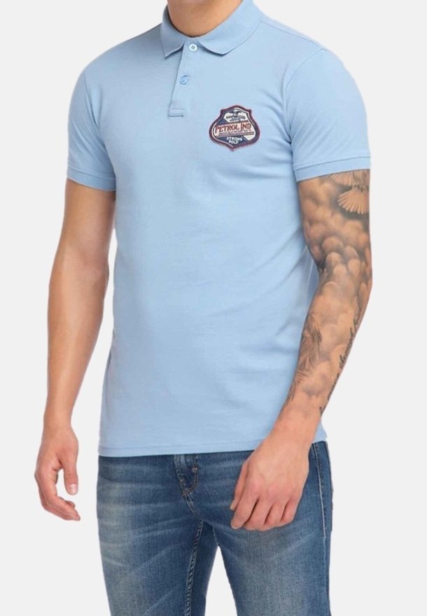 Homme portant un polo bleu clair avec un écusson sur la poitrine et un jean bleu, montrant une manche de tatouage détaillée sur son bras droit.