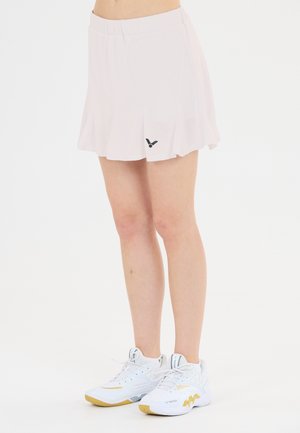 Kvinde iført hvid plisseret tennisnederdel og hvide sportssko med gule accenter, stående på en ensfarvet hvid baggrund.