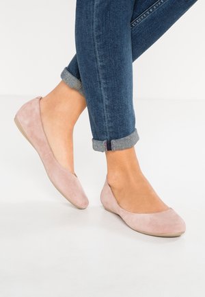 Pieds portant un jean bleu retroussé et des ballerines en daim rose clair sur un sol blanc, un pied légèrement levé.