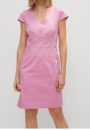 Femme portant une robe rose ajustée, longueur genoux, avec des manches courtes et un décolleté en V, sur un fond clair uni.