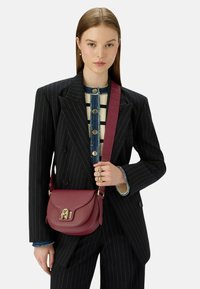 Žena v černém pruhovaném obleku a pruhovaném svetr s knoflíky, držící vínově červenou koženou crossbody tašku se zlatým uzávěrem.