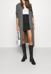 Šedý dlouhý kardigan přes bílý top, spárovaný s černou džínovou mini sukni. Doplňky zahrnují texturovanou bílou tašku a černé vysoké kozačky.