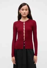 Burgundi spalvos raukšlėta megztinė su apvalia iškirpte, su mygtukų užsegimu priekyje, ovaliais išpjovimais ir dekoratyviais mygtukais. Ilgos rankovės.