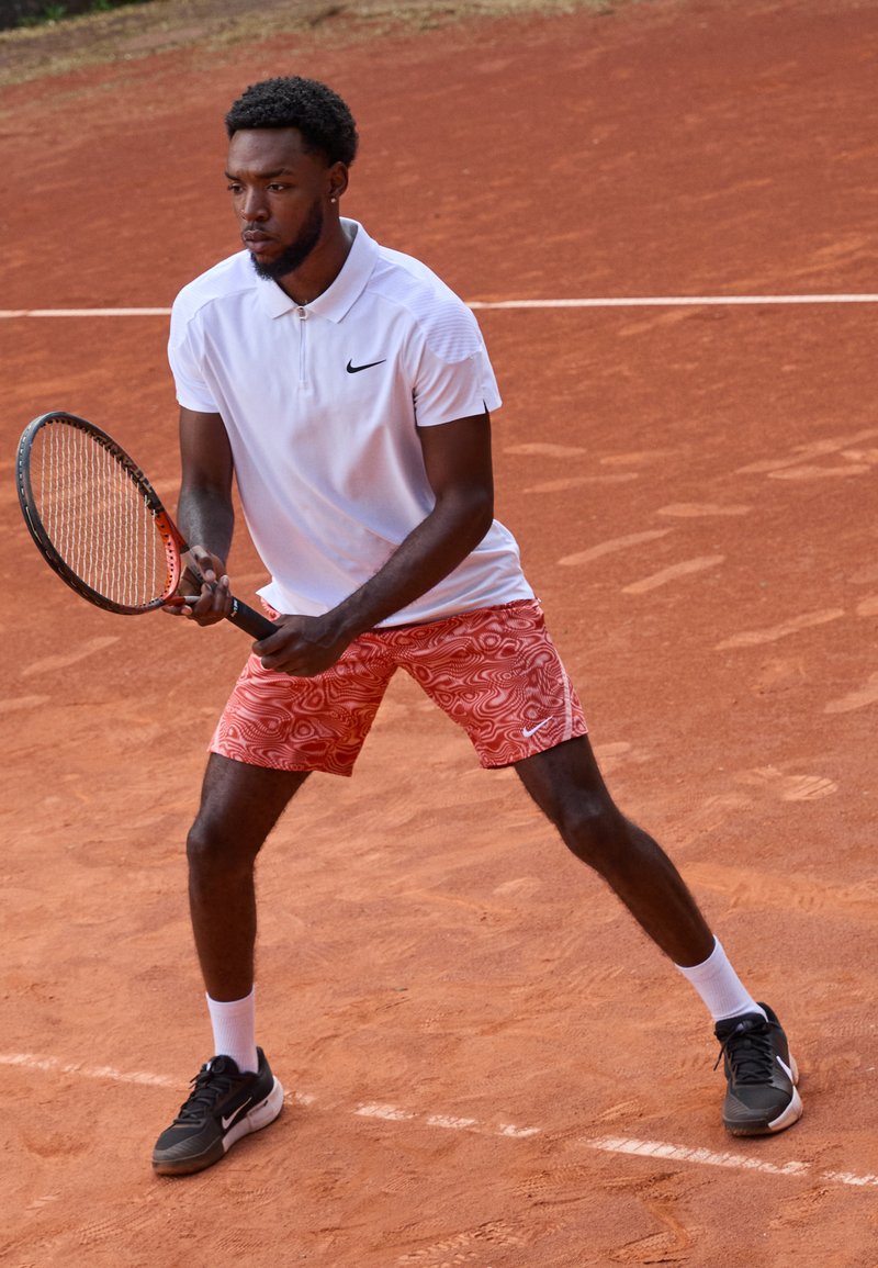 Biała koszulka polo z logo Nike, czerwone spodenki w wzory, czarno-białe buty tenisowe, trzymając rakietę tenisową na korcie ziemnym.