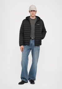 Jachetă puffer neagră cu glugă, pulover gri cu model dedesubt, blugi evazați bleumarin deschis și pantofi negri. Capac gri deschis cu logo.