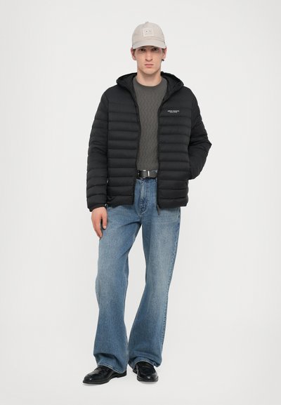 Doudoune noire avec capuche, pull gris à motifs en dessous, jean évasé bleu clair et chaussures noires. Casquette gris clair avec logo.