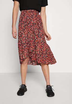 Kvinde iført et knælængde, rødt blomsterprint slå-om-nederdel med sorte sneakers og en sort kortærmet top mod en ensfarvet baggrund.