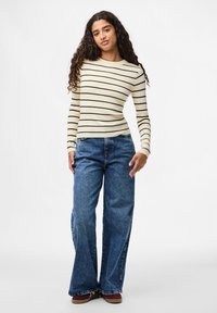 Πλεκτό πουλόβερ με ριμπ δομή σε κρεμ απόχρωση με μαύρες ρίγες, συνδυασμένο με φαρδιά τζιν παντελόνια σε μπλε χρώμα. Διαθέτει μακριά μανίκια και στρογγυλή λαιμόκοψη.