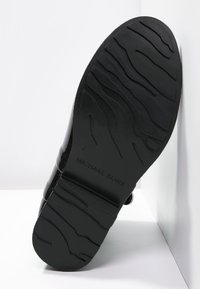Semelle noire d'une chaussure Michael Kors avec un motif de semelle texturé en vagues, présentée en angle contre un fond de coin blanc.