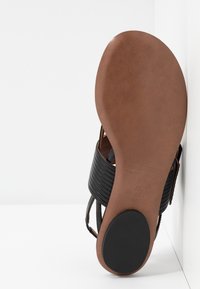 Sandalo in pelle nera con design del cinturino rigato, suola in finta pelle marrone, profilo piatto e fondo liscio con grip circolare in gomma nera.