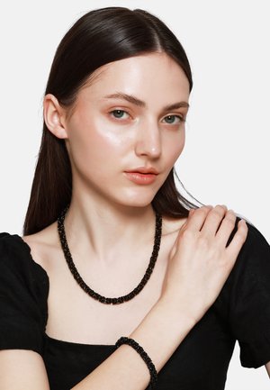 Schwarzes Perlenketten- und Armband-Set. Die Halskette besteht aus glatten, runden Perlen. Das Model trägt ein schwarzes Oberteil vor einem neutralen Hintergrund.