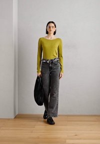 Grønn langermet topp, svart høy midje jeans, kraftige svarte sko, overdimensjonert svart veske og klare solbriller, foran en nøytral bakgrunn.