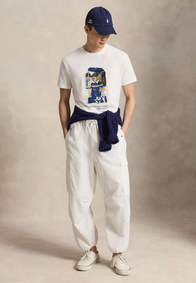 Άνδρας που φοράει λευκό graphic t-shirt, λευκό cargo παντελόνι, λευκά αθλητικά παπούτσια, μπλε σκούφο, με μπλε πουλόβερ δεμένο γύρω από τη μέση, στέκεται σε ουδέτερο φόντο.