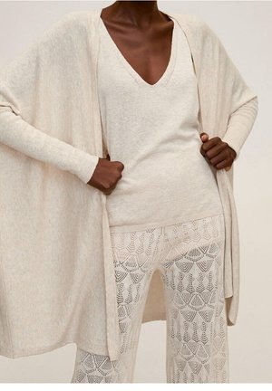 Dunkelhäutige Person trägt beige V-Ausschnitt Top, offene Strickjacke und gemusterte Strickhose, Hände halten die Kanten der Strickjacke.