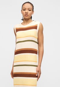 Γυναίκα με πλεξουδωτά μαλλιά που φοράει αμάνικο πλεκτό φόρεμα με οριζόντιες ρίγες σε εκρού, καφέ, κίτρινο, πορτοκαλί και ελιά.