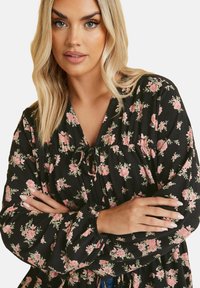 Blusa nera a fiori con rose rosa, caratterizzata da scollatura a V, dettaglio con lacci e maniche lunghe con polsini elasticizzati, realizzata in tessuto leggero.