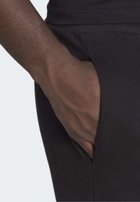 Tmavou pletí ruka zasunutá do boční kapsy černých žebrovaných kalhot, zobrazená na světlém pozadí.