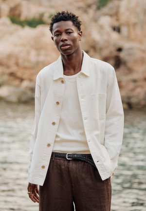 Fiatal férfi fehér gombos ingben, krémszínű pulóverben és barna nadrágban, víz közelében, sziklás háttér előtt áll.