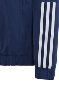 Dunkelblaue Jacke mit elastischen Bündchen, versehen mit drei weißen vertikalen Streifen an den Ärmeln. Hergestellt aus glattem, leichtem Stoff.