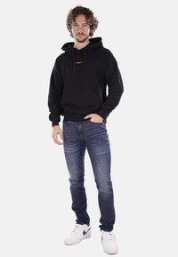 Felpa nera con cappuccio e tasca frontale, con logo arancione. Abbinata a jeans in denim blu e sneakers bianche.