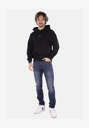 Felpa nera con cappuccio e tasca frontale, con logo arancione. Abbinata a jeans in denim blu e sneakers bianche.